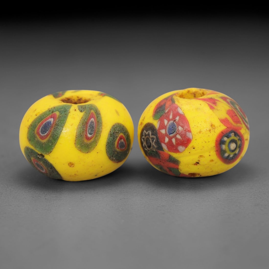 A Pair of Venetian Millefiori Glass Beads, 19th-20th Century, 52AD: Fabricated from opaque yellow glass, each bead is formed as a short cylinder with a central drilled perforation. The surface is decorated with characteristic millefiori canes: one bead displays eye-li