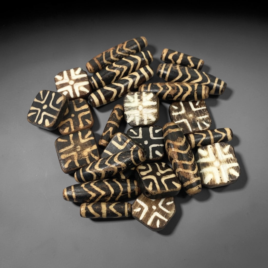 A Group of Burmese Pumtek Wood Beads, 19th-20th Century, 11FT: The group consists of twenty-five beads, carved from wood and treated with a dark surface, overlaid with white geometric patterns. The beads appear in two primary forms: cylindrical and square or rect