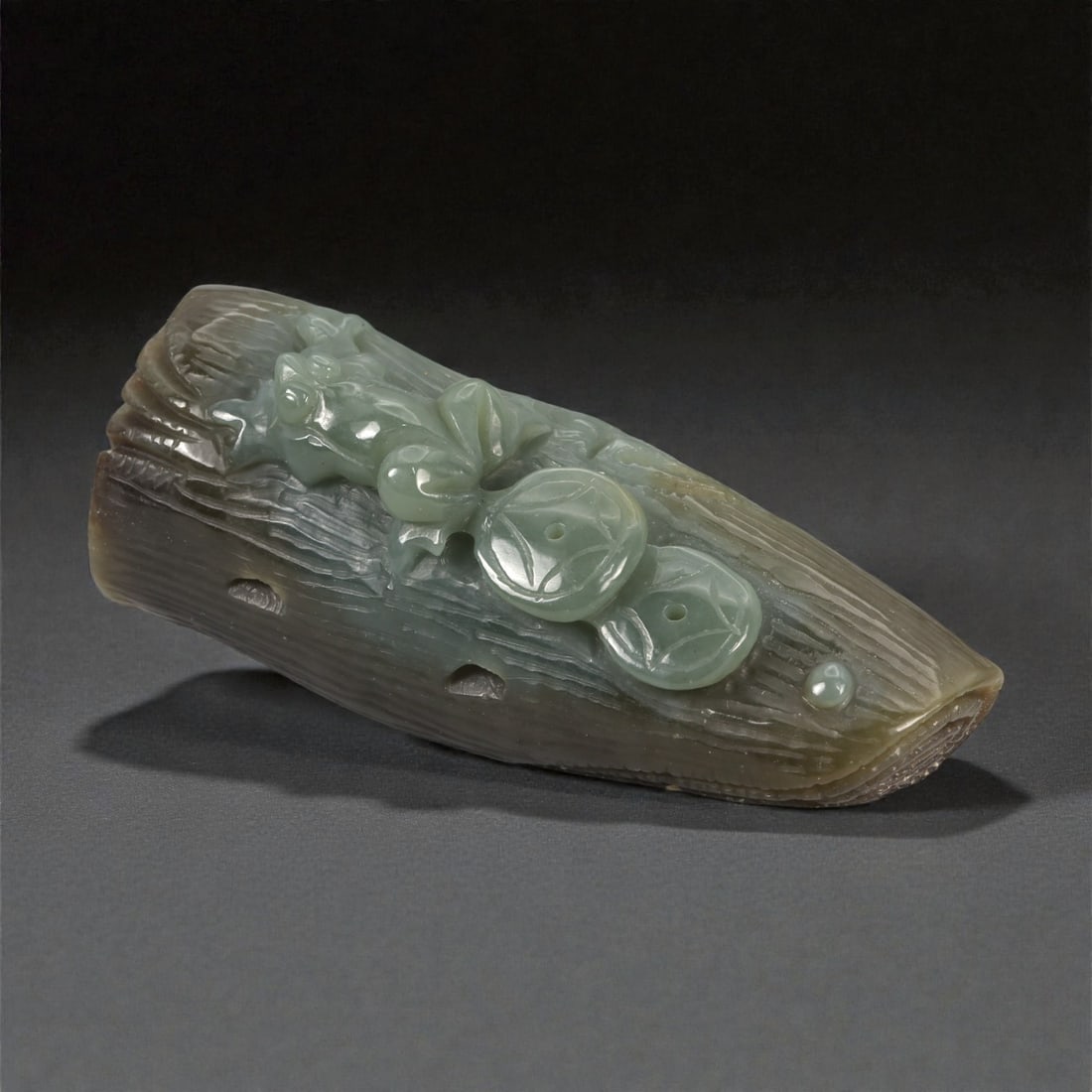 A Chinese Jade Ruyi Fungus and Coin Carving, 20th Century, 61YA: Carved from grey-green jade with areas of darker brown and olive-toned inclusions, the object is worked as an elongated, gently tapering form resembling a section of wood or branch. The upper surface