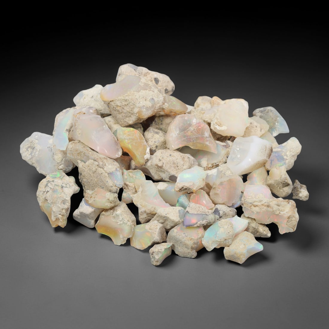 An Ethiopian Welo Opal Rough Lot, 50.75g, 54ZH: A parcel of rough Welo opal, with individual pieces ranging from 1-2.5 cm in size. The specimens exhibit a milky to translucent base, with visible play-of-color displaying flashes of blue, green, yell