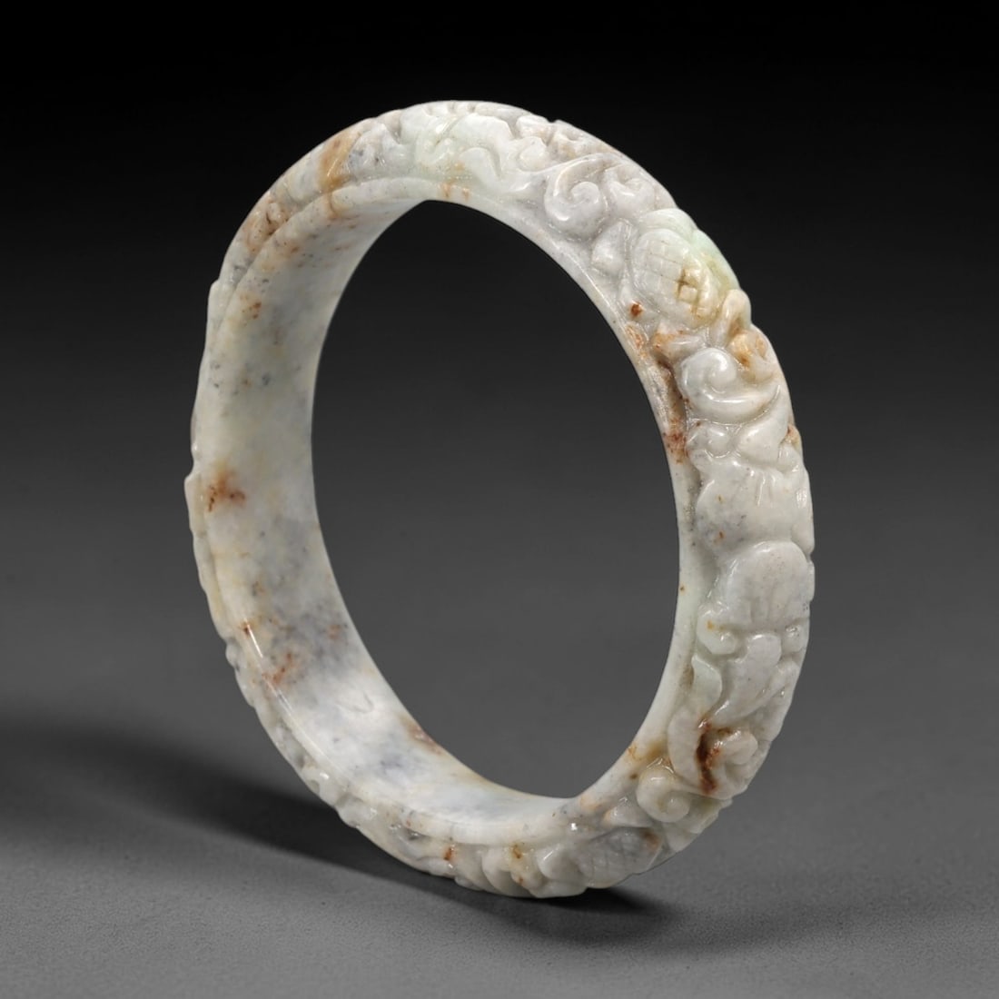 A Chinese Carved Jadeite Jade Bangle Bracelet, 20th Century, 65ZL: A fine jadeite bangle of flattened circular form, masterfully carved in relief with a continuous band of scrolling foliate and auspicious lingzhi motifs. The stone exhibits a translucent pale white ba