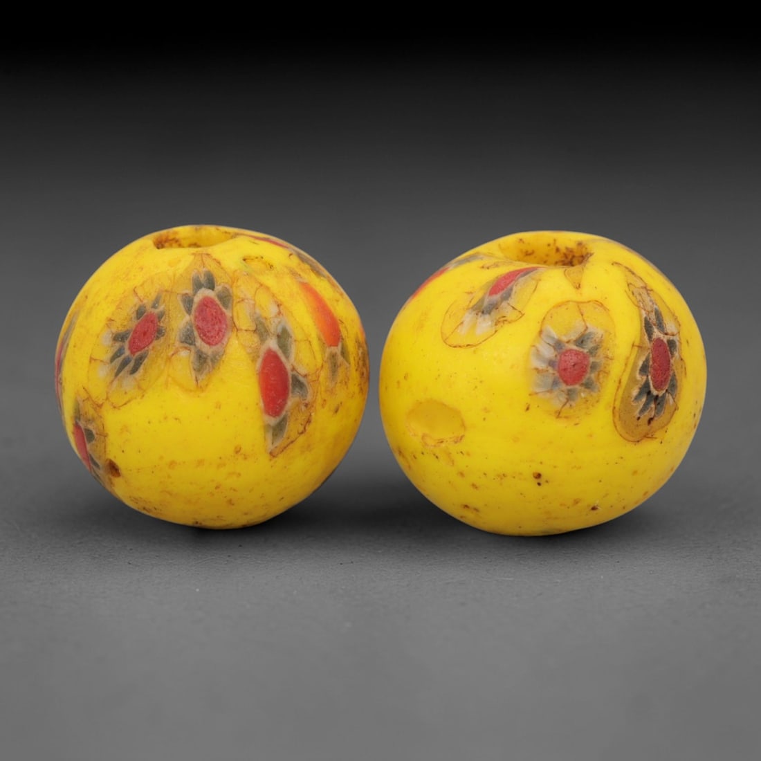 A Pair of Venetian Millefiori Glass Beads, 19th-20th Century, 56AD: Fabricated from opaque yellow glass, each bead is formed as a sphere with a central drilled perforation. The surface is decorated with characteristic millefiori canes, featuring floral and eye-like mo