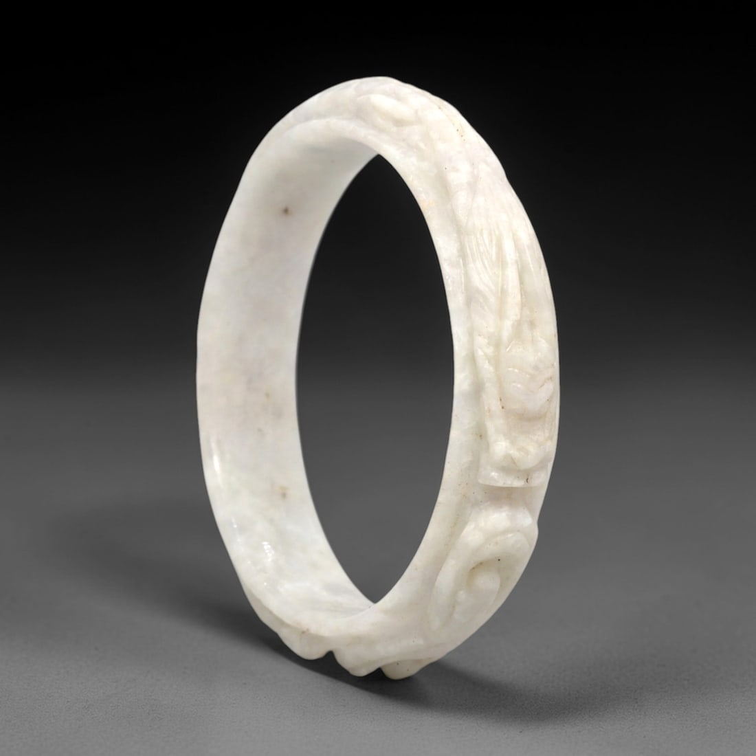 A Chinese Jadeite Jade Bangle Bracelet, 20th Century, 28FS: Carved from pale milky-white jadeite jade with minor natural inclusions, the bangle is formed as a continuous circular band. The exterior surface is decorated with raised relief carving depicting a co