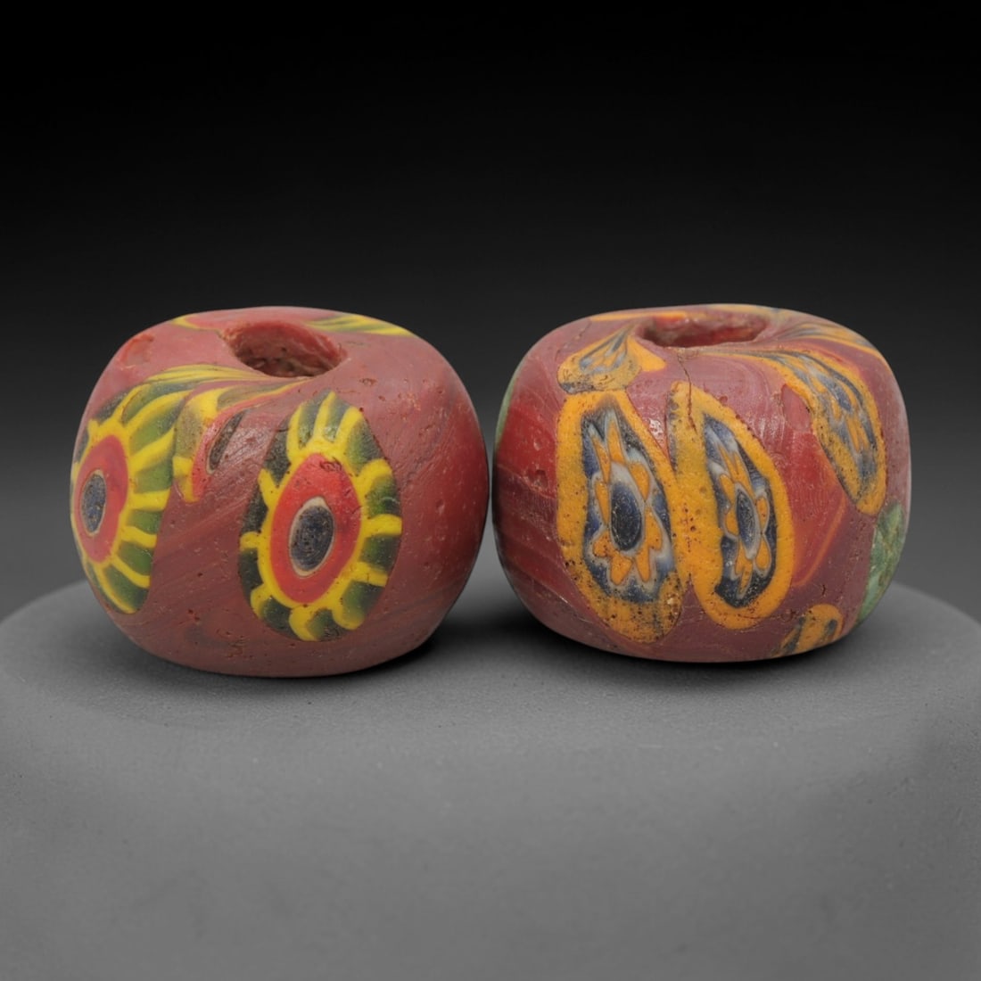 A Pair of Venetian Millefiori Glass Beads, 19th-20th Century, 74AD: Fabricated from opaque red glass, each bead is formed as a short cylinder with a central drilled perforation. The surface is decorated with characteristic millefiori canes: one bead displays circular