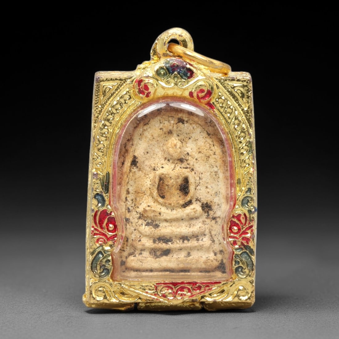 A Thai Phra Somdet Clay Amulet in a Gilded Enamel Case, 20th Century, 19ZK: This rectangular Phra Somdet amulet is crafted from sacred beige clay and encased in an ornate gilded metal case with a suspension loop at the apex. The obverse of the case is elaborately decorated wi