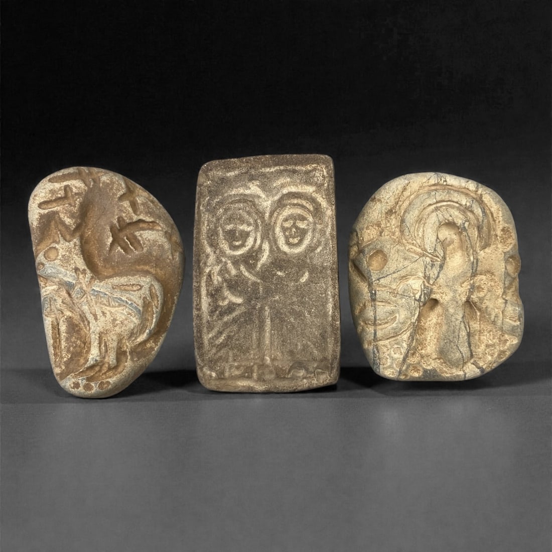 A Group of Three Christian Period Stone Carving Reliefs, 4th-7th Century, 62FM: Comprising three stone carving reliefs, each formed from naturally shaped or cut stone with visible surface fissures and tonal variation. The leftmost relief depicts a stylized quadruped, likely a dee