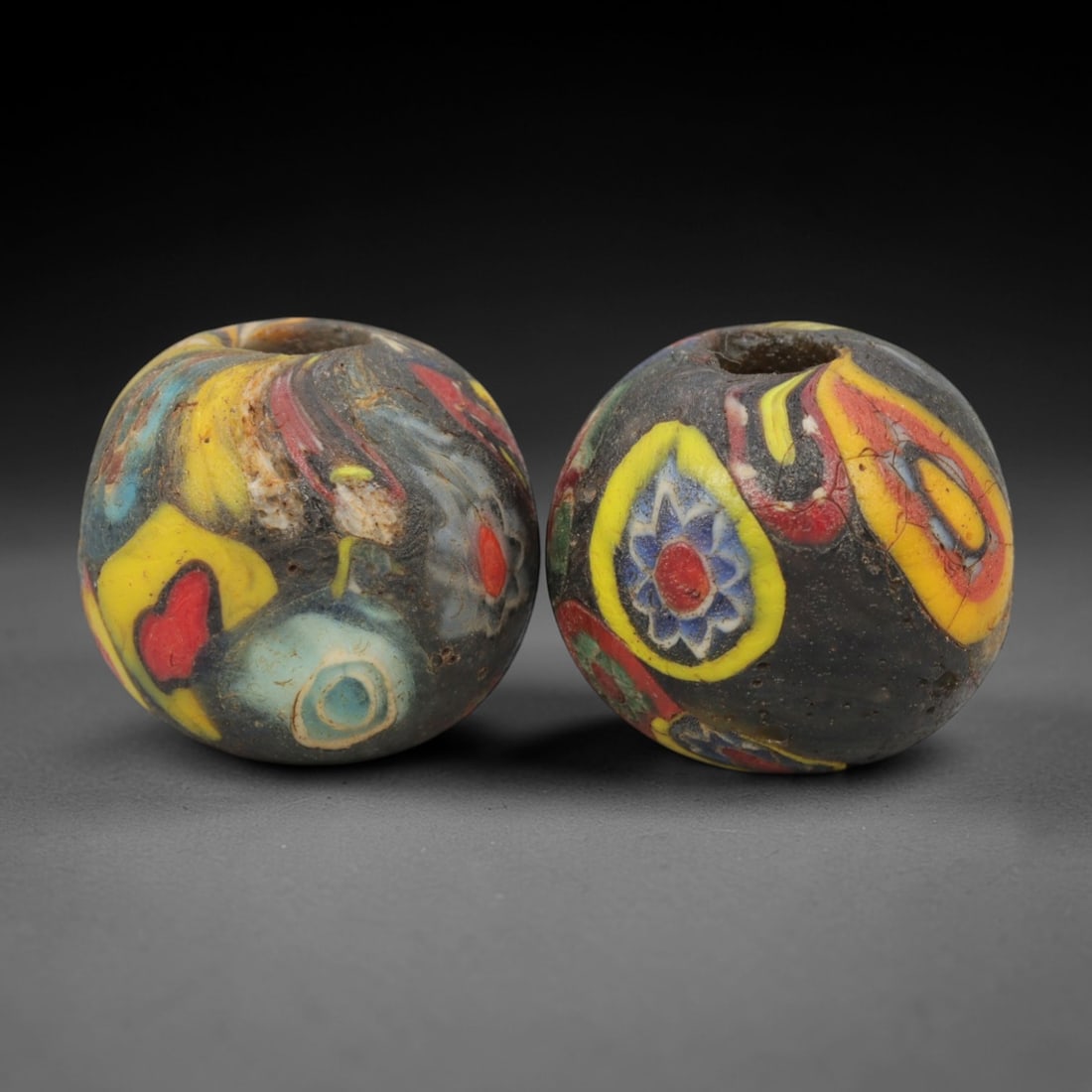 A Pair of Venetian Millefiori Glass Beads, 19th-20th Century, 54AD: Fabricated from opaque dark glass, each bead is formed as a sphere with a central drilled perforation. The surface is decorated with characteristic millefiori canes, featuring floral and abstract moti