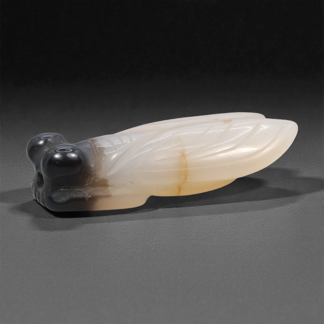 A Chinese Agate Cicada Ornament, 20th Century, 95FH: Carved from translucent agate with graduated coloration ranging from milky white to smoky gray and pale amber tones, the ornament is modeled in the form of a cicada shown in repose. The head is distin