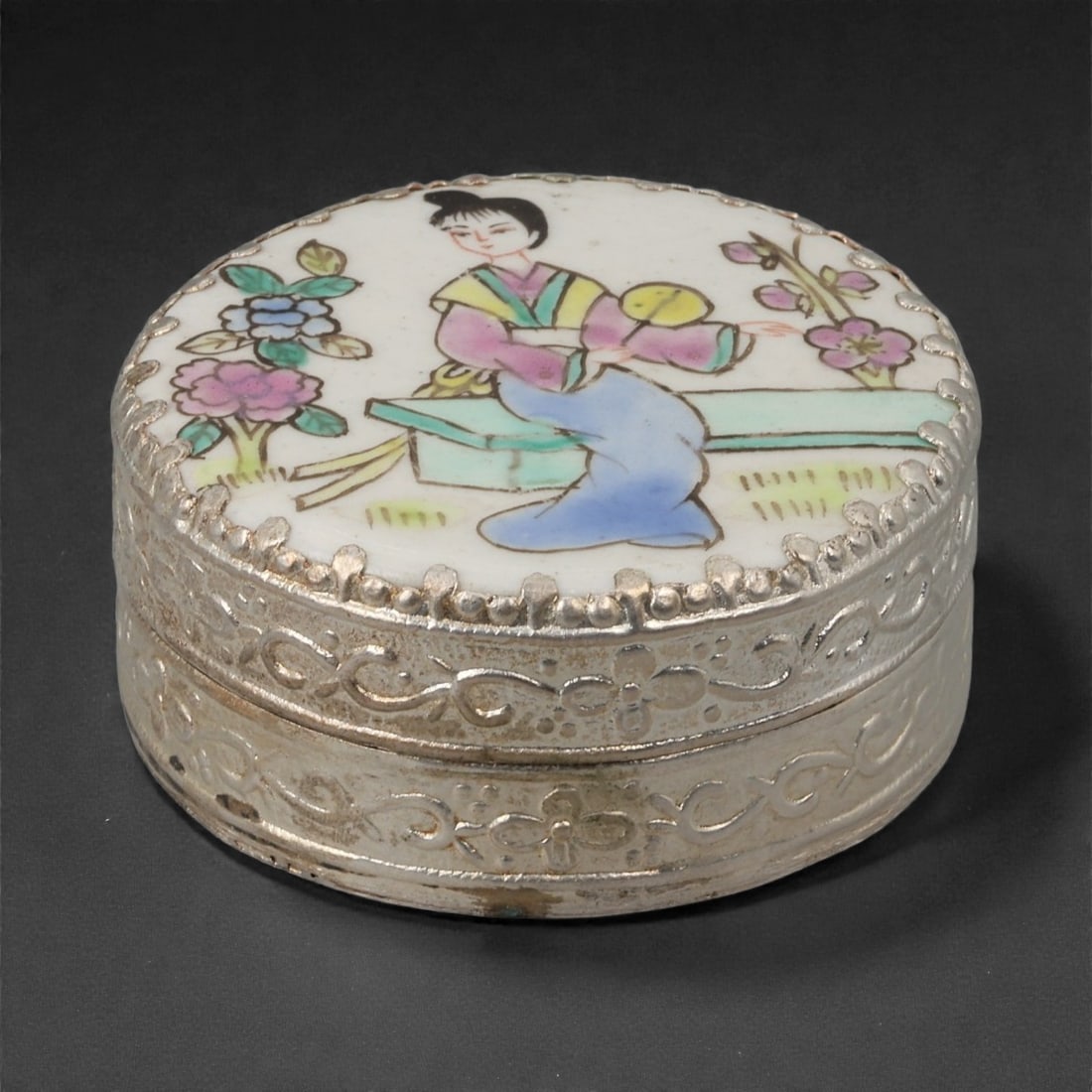 A Chinese Silvered Porcelain Trinket Box, 20th Century, 82FG: Constructed with a circular porcelain lid set within a silvered metal mount, the trinket box is formed as a shallow cylindrical container with a fitted cover. The porcelain lid is decorated in polychr