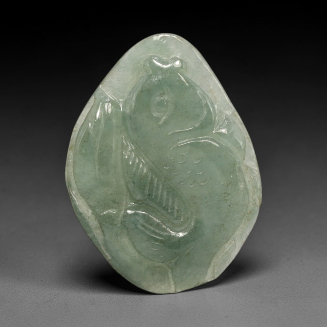 A Chinese Jadeite Jade Fish Pendant, 20th Century, 70FR: Carved from translucent pale green jadeite jade with subtle natural tonal variations, the pendant is shaped as a polished pebble form. It depicts a fish rendered in low relief, with incised lines defi
