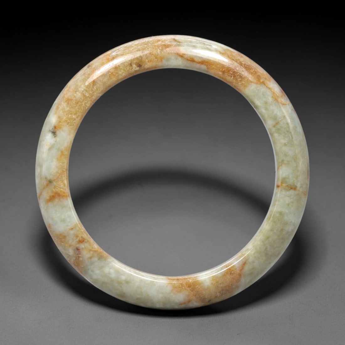 A Burmese Jadeite Jade Bangle, 20th Century, 7FS: A Burmese jadeite jade bangle, with a pale greenish-white matrix and prominent russet-brown mineral inclusions. The bangle is of circular form, with a smooth, polished surface revealing the internal c