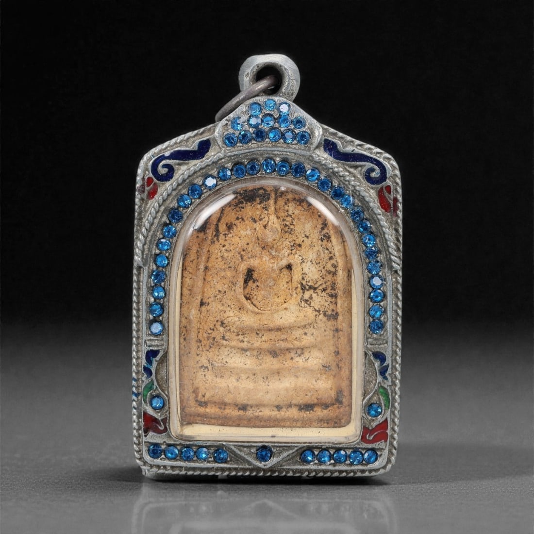 A Thai Buddha Amulet with Enameled Metal Frame, 20th Century, 39ZH: The amulet is of rectangular form with a stepped upper edge. The obverse features a molded seated Buddha figure in meditative pose (dhyana mudra), rendered in a light beige composite material with inc