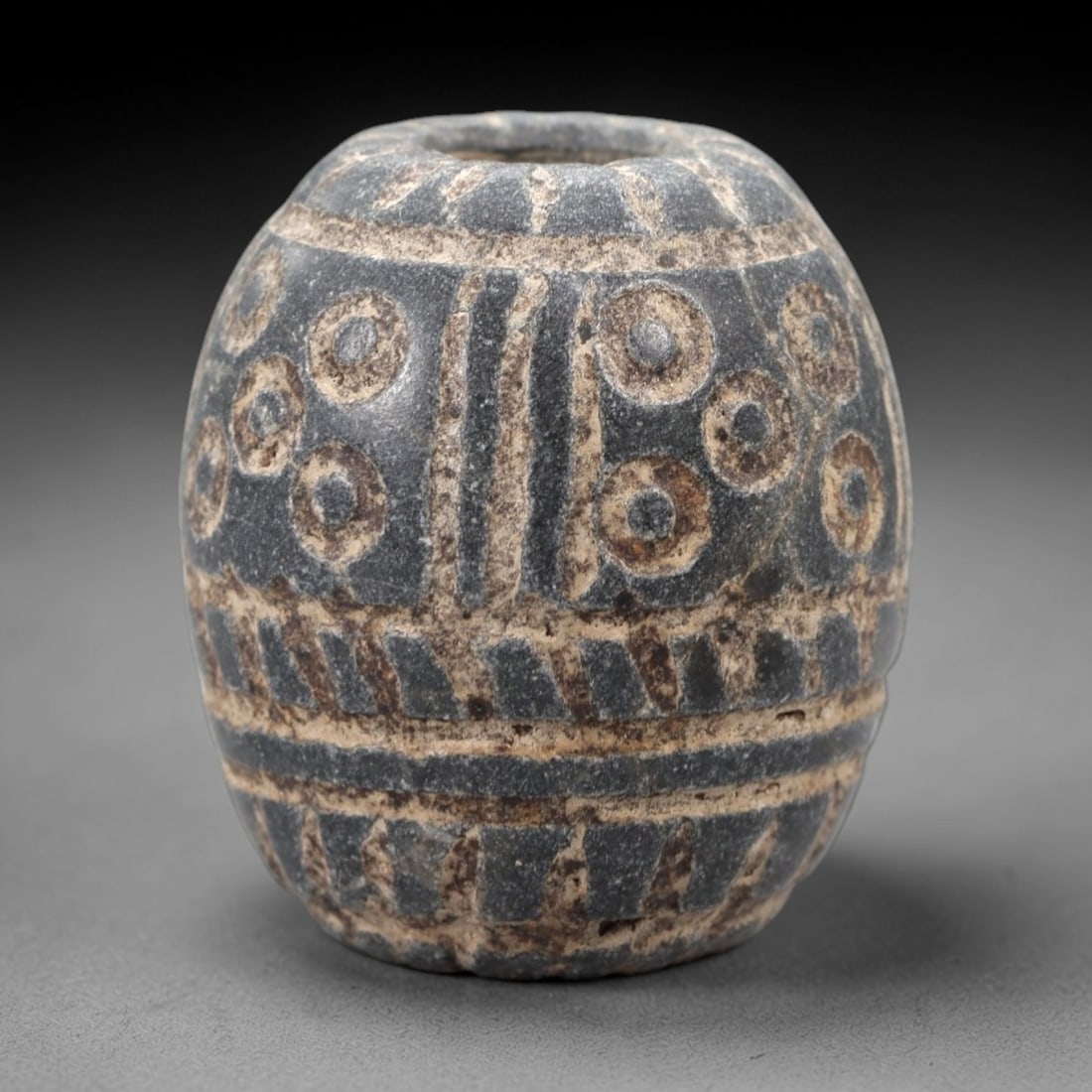 A Near Eastern Schist Barrel Bead, Circa 1st Millennium BCE, 10FT: Carved from dark grey schist with a fine granular texture, the object is a barrel-shaped bead featuring a central vertical perforation for suspension. The exterior is ornamented with incised geometric