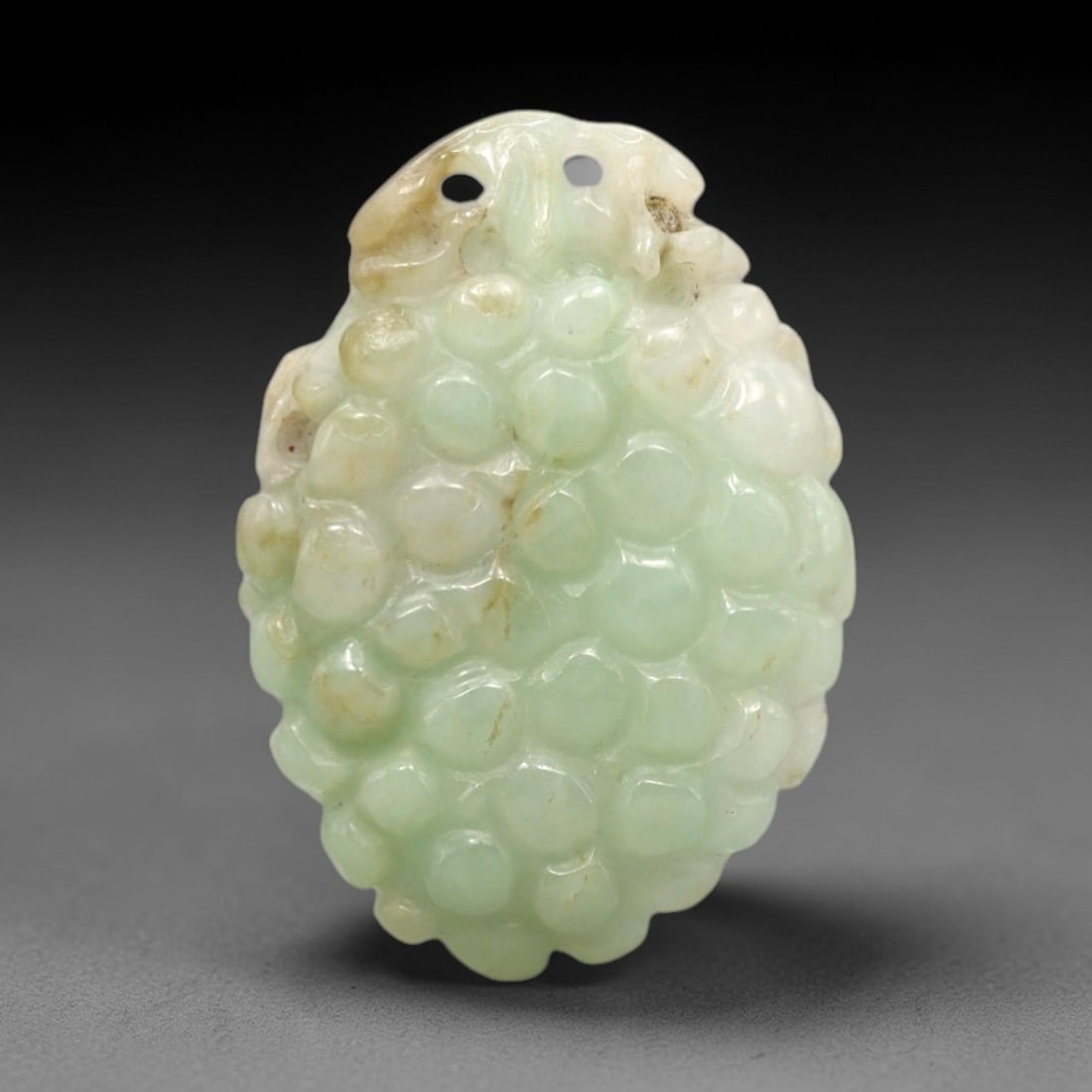 A Chinese Jadeite Jade Grape Pendant, 20th Century, 71FR: Carved from pale green jadeite jade with natural tonal variation and scattered russet inclusions, the pendant is shaped as an oval form with two top suspension holes. A cluster of grapes is rendered i