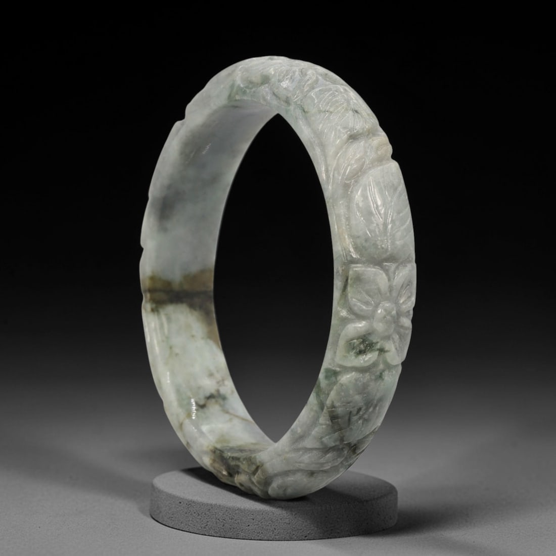 A Chinese Carved Jadeite Jade Bangle Bracelet, 20th Century, 45ZL: A fine jadeite bangle of flattened circular form, masterfully carved in relief with a continuous band of floral blossoms and scrolling foliate motifs. The stone exhibits a translucent pale greenish-gr