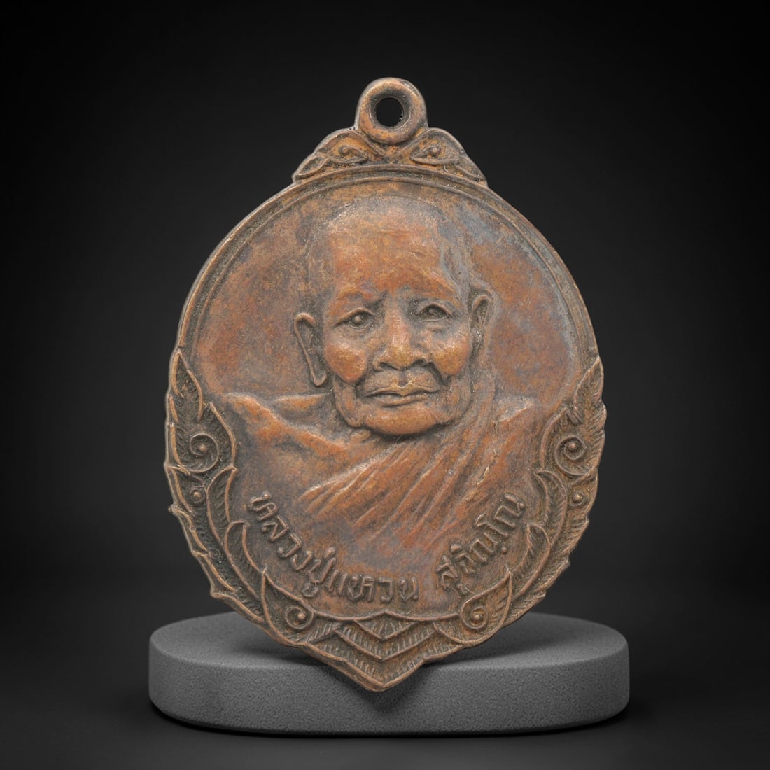 A Thai Copper Alloy Buddhist Amulet, Late 20th Century, 94FP: A circular copper alloy amulet with a pointed base, measuring 4 x 2.8 cm. The obverse depicts a bust of a seated Buddhist monk in relief, framed by ornate scrollwork, with Thai script inscriptions bel