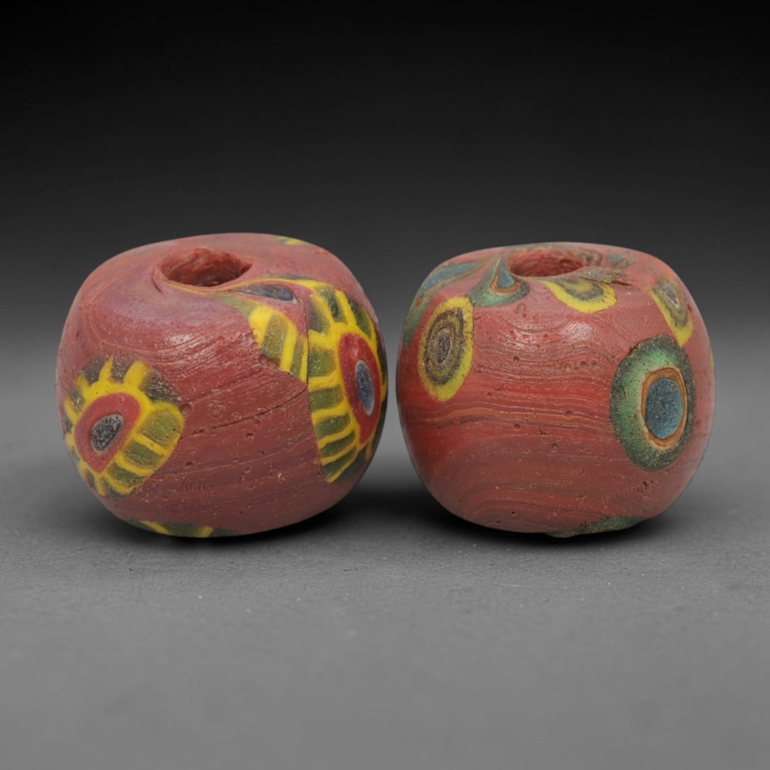 A Pair of Venetian Millefiori Glass Beads, 19th-20th Century, 44AD: Fabricated from opaque red glass, each bead is formed as a short cylinder with a central drilled perforation. The surface is decorated with characteristic millefiori canes: one bead displays circular