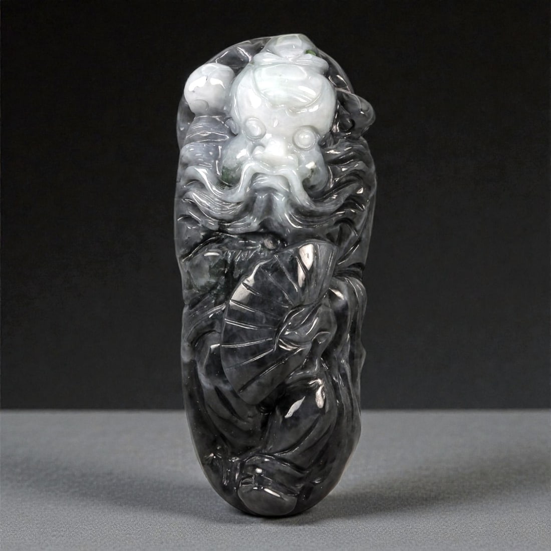 A Chinese Jadeite Jade Figural Pendant, 20th Century, 32FB: Carved from dark grey to black jadeite with areas of milky white translucency concentrated in the upper section, the pendant is worked in high relief as a standing male figure holding an open fan. The
