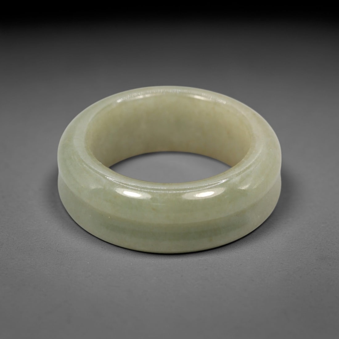 A Chinese Jadeite Jade Ring, 20th Century, 13XN: Carved from greenish jadeite jade, the ring is formed as a thick, circular band with a smooth, highly polished surface. The form features a wide, gently convex outer profile and a concave inner surfac