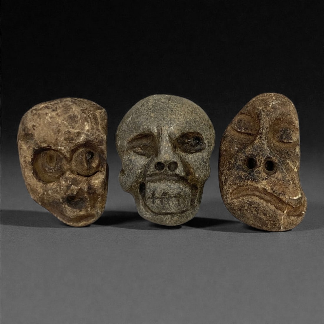 A Group of Three Medieval Stone Maskette Amulets, 10th-14th Century, 48FP: The group comprises three stone maskette amulets, each carved from distinct stone types with varied surface treatments. The first is a light brown stone mask with an irregular form, featuring recessed