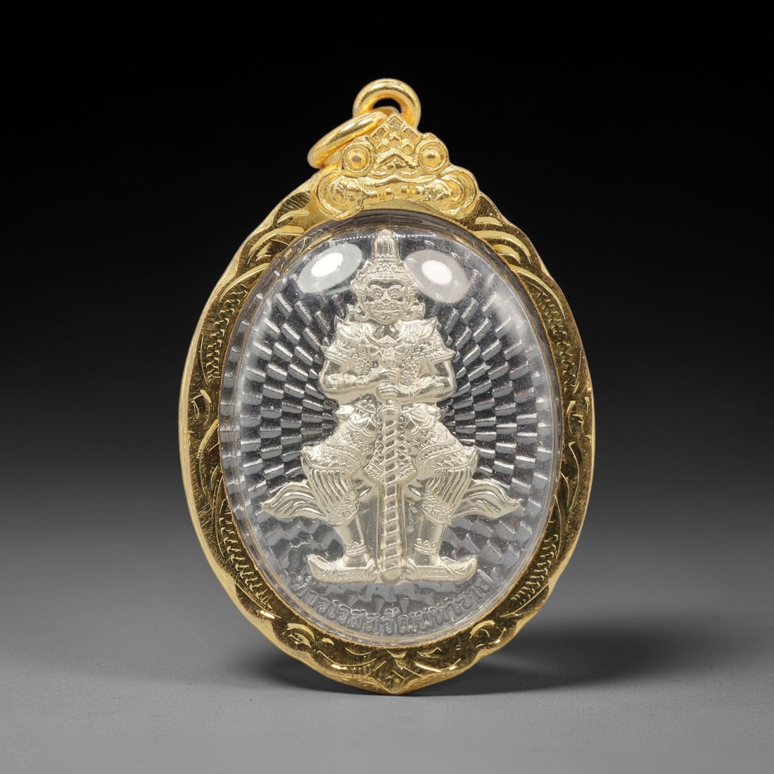 A Thai Guardian Deity Amulet in Gold Frame, Late 20th Century, 4FR: An oval-shaped amulet, measuring 5 x 3.5 cm, set within an ornate gold frame with a suspension loop. The obverse depicts a seated guardian deity in gold and silver relief, holding a staff, set against