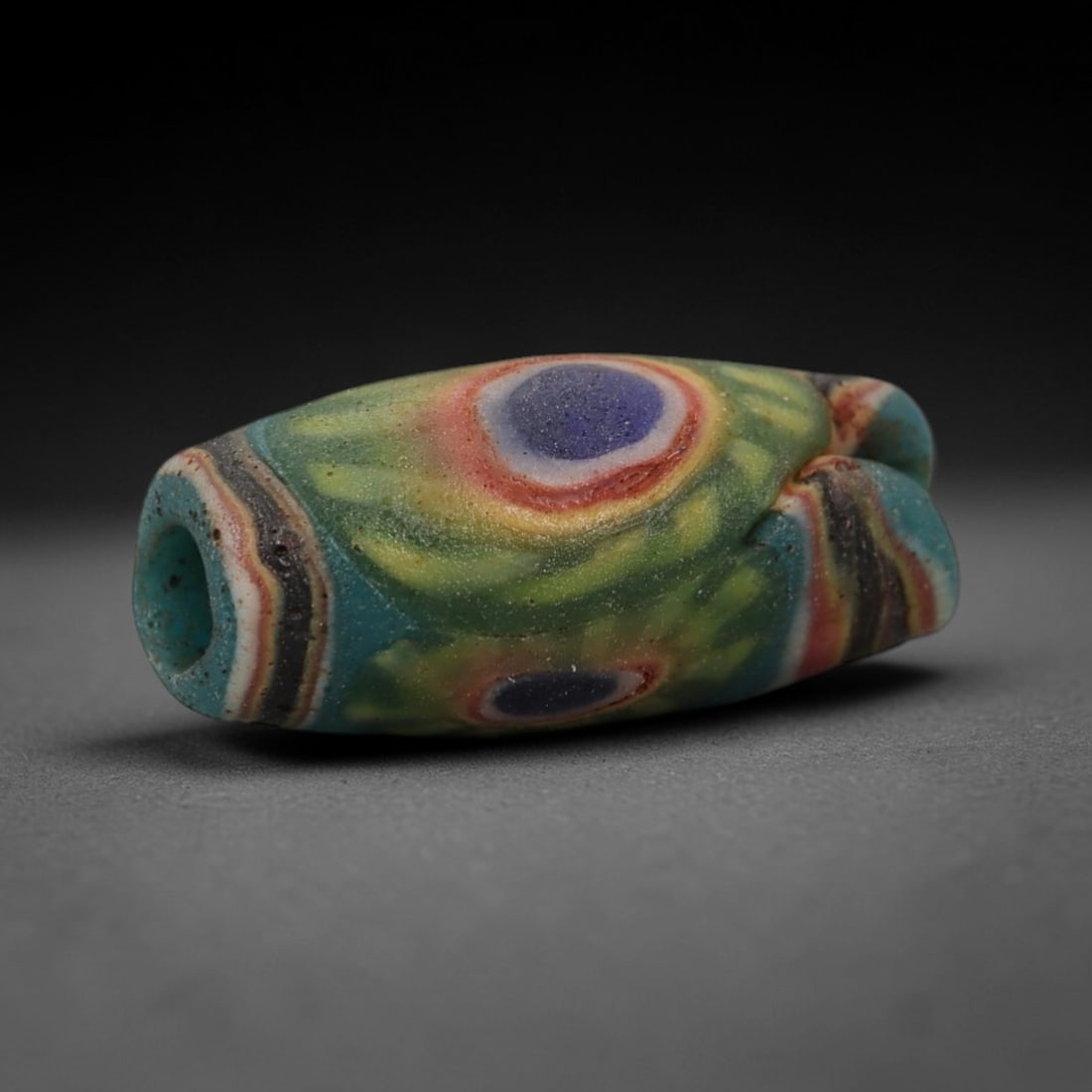 A Sino-Himalayan Glass Eye Bead, 18th-19th Century, 1FW: Formed from translucent blue-green glass with swirling yellow and pale green filamentary inclusions characteristic of mosaic glass working, the bead is barrel-shaped with a central perforation for sus