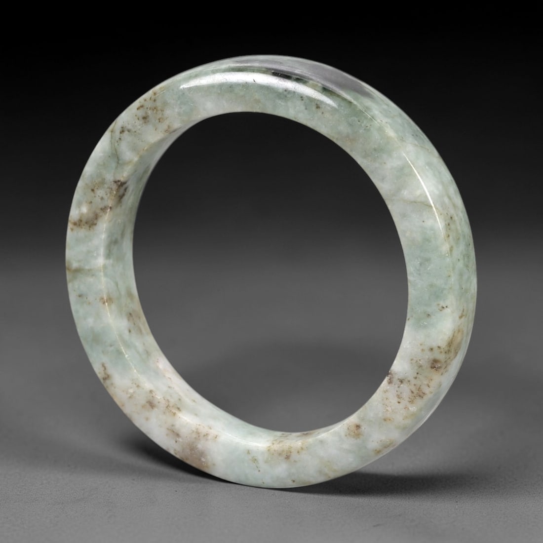 A Chinese Jadeite Jade Bangle Bracelet, 20th Century, 87ZK: Carved from jadeite jade with a pale green base and scattered brownish inclusions, the bangle is formed as a continuous circular band with a rounded cross-section. The surface is smoothly polished, wi