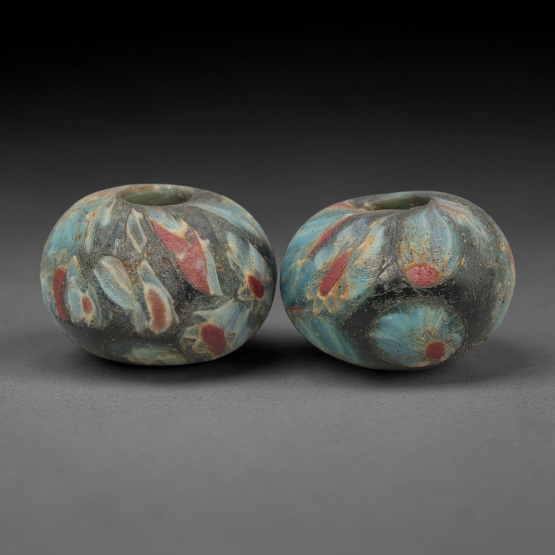 A Pair of Venetian Millefiori Glass Beads, 19th-20th Century, 58AD: Fabricated from opaque red glass, each bead is formed as a short cylinder with a central drilled perforation. The surface is decorated with characteristic millefiori canes, featuring eye-like motifs w