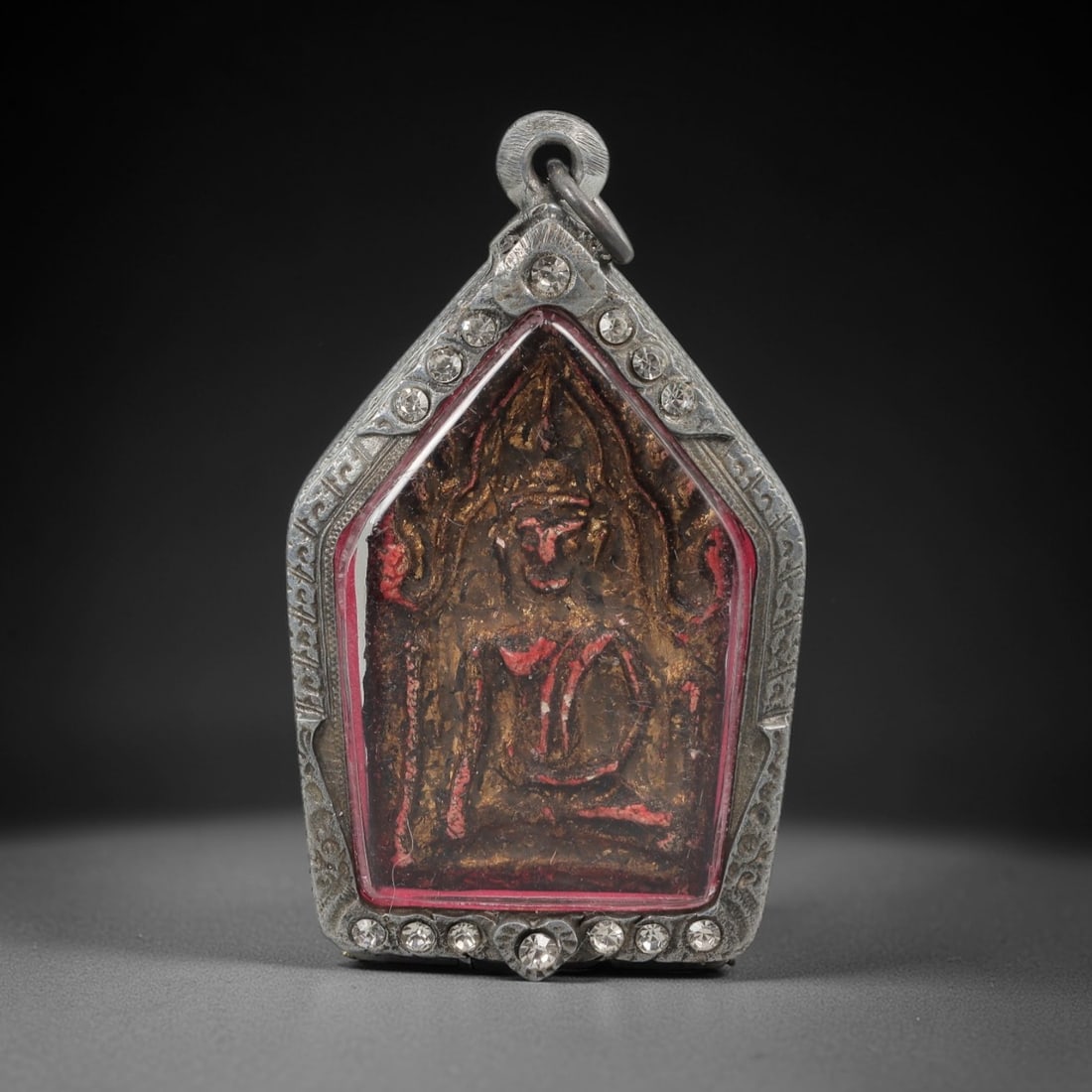 A Thai Phra Khun Paen Amulet in a Silver Repousse Frame, 20th Century, 81FT: The amulet features a tapering metal plaque depicting Phra Khun Paen in a seated meditative pose, rendered in a textured alloy with layered patinas of brown, red, and gilded toning characteristic of s