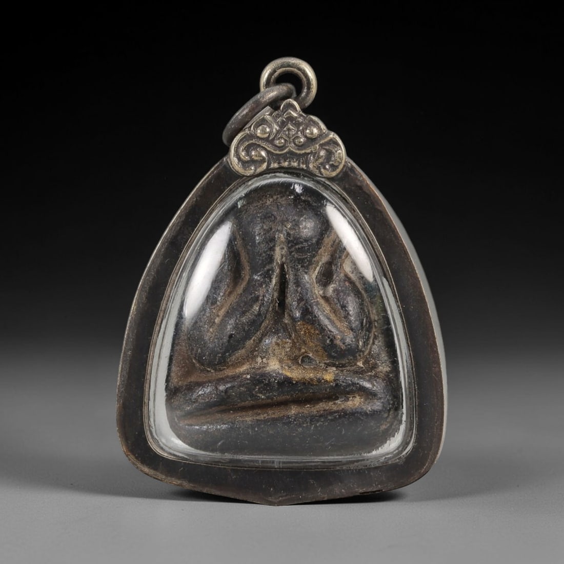 A Thai Phra Pidta Amulet, 20th Century, 84FT: The amulet depicts Phra Pidta, the Buddha of protection, cast in a dark, patinated metal with visible surface wear and residual mineral deposits. The figure is shown seated in a meditative pose, with