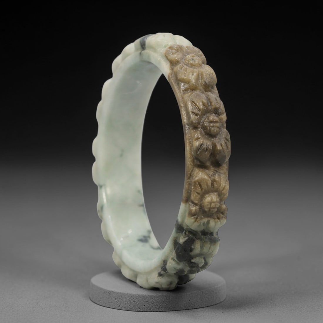 A Chinese Carved Jadeite Jade Bangle Bracelet, 20th Century, 71ZL: A fine jadeite bangle of flattened circular form, masterfully carved in relief with a continuous band of chrysanthemum blossoms. The stone exhibits a striking variegated palette, with a pale milky-gre