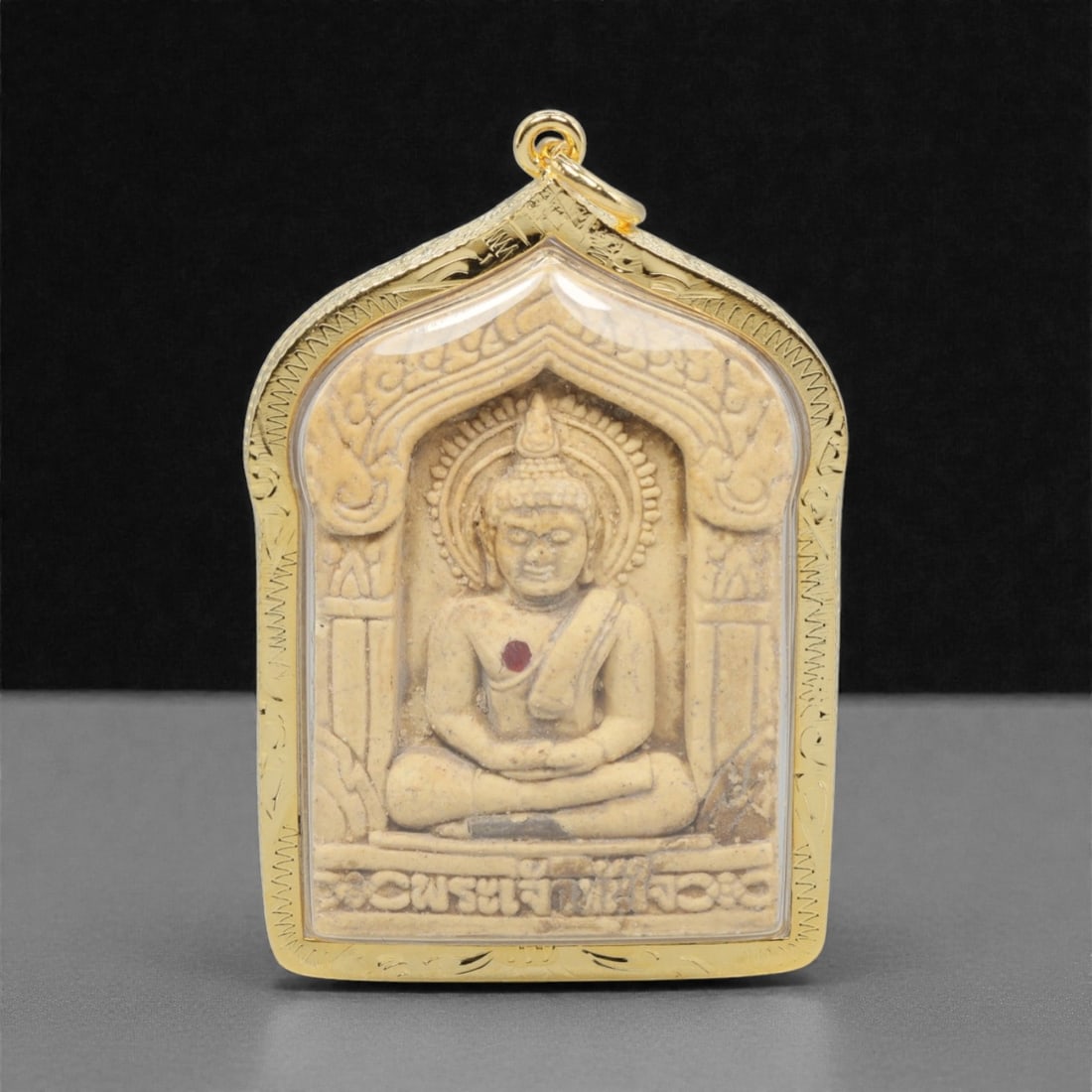 A Thai Khmer-Style Buddha Amulet Pendant, 20th Century, 21FP: This pendant features a light beige composite amulet core, encased in an ornate gilded metal frame with a transparent protective cover, measuring 6.1 x 4 cm (2 3/8 x 1 9/16 in.). The obverse displays