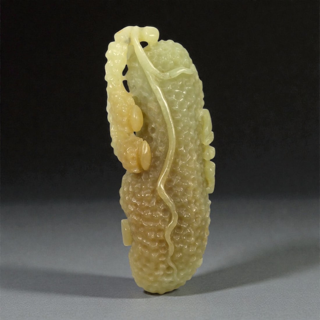 A Chinese Nephrite Jade Fruit-Form Pendant, 20th Century, 70FA: Carved from pale celadon nephrite jade with areas of warm yellow-brown tonal variation and moderate translucency, the pendant is modeled in the form of an elongated fruit. The surface is densely worke