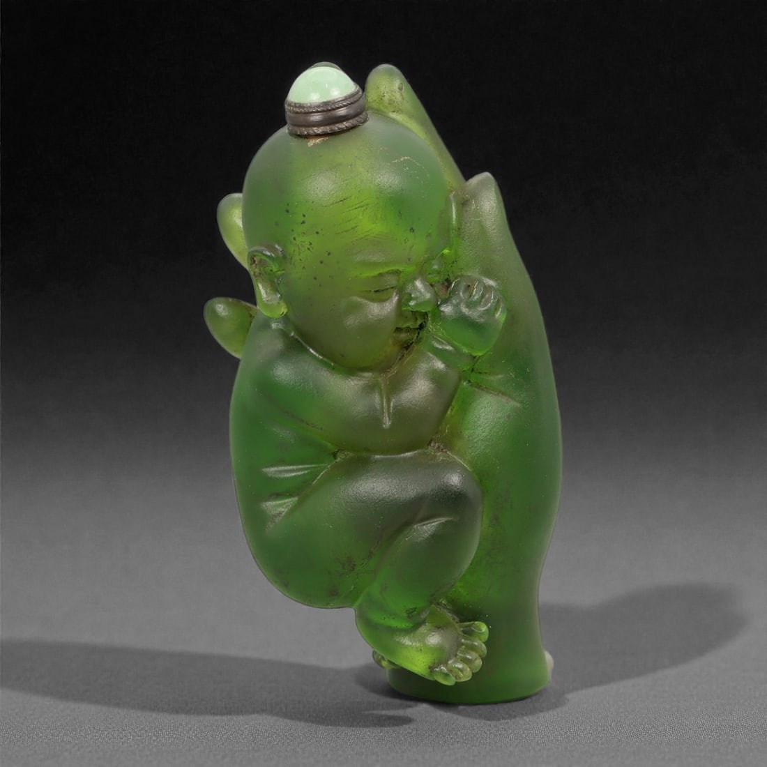 A Chinese Glass Figural Snuff Bottle, 20th Century, 71FG, 71FG: Molded from translucent green glass with darker and lighter tonal variations, the snuff bottle is formed as a crouching infant figure shown in high relief against an integrated backing plane. The chil
