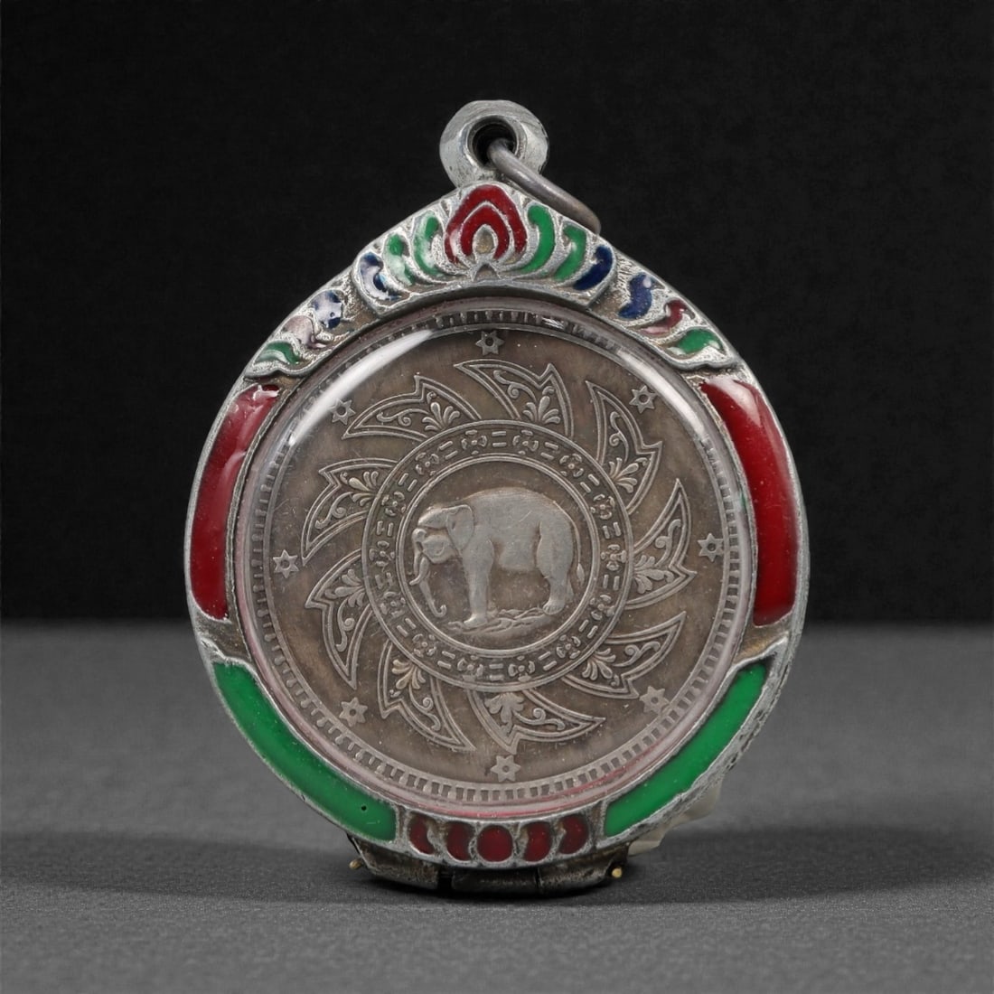 A Thai Silvered Metal Medallion in Enameled Silvered Metal Frame, 20th Century, 53FN: The medallion is encased in a circular silvered metal frame with a suspension loop at the apex. The obverse frame is embellished with applied red, green, and blue enamel inlay within chased borders; t