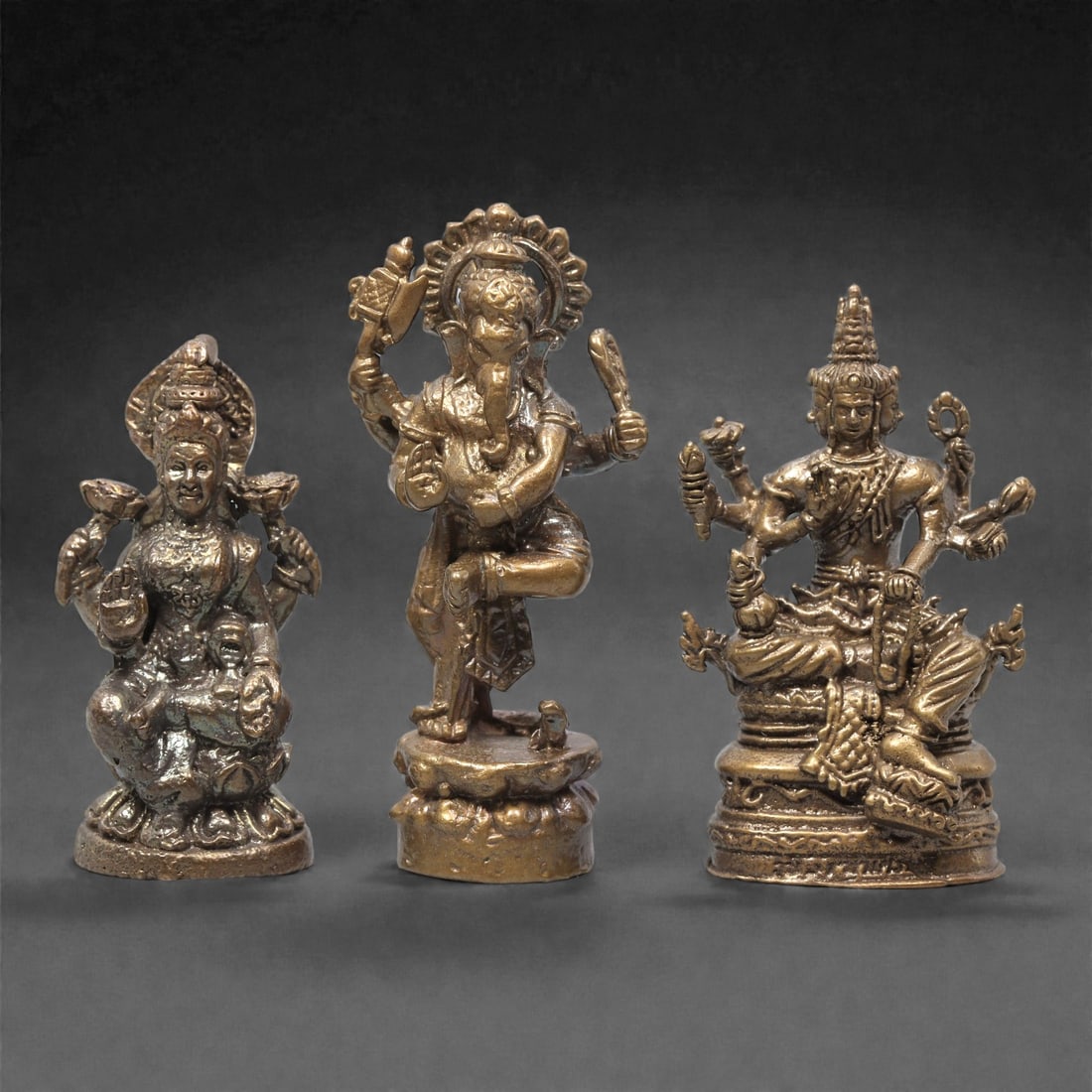 A Set of Three Thai Brass Hindu Deity Figures, 20th Century, 70ZG: Cast from brass with a dark, aged patina, the set comprises three distinct Hindu deity figures. The left figure is a seated multi-armed goddess, holding symbolic attributes and a child figure on her l