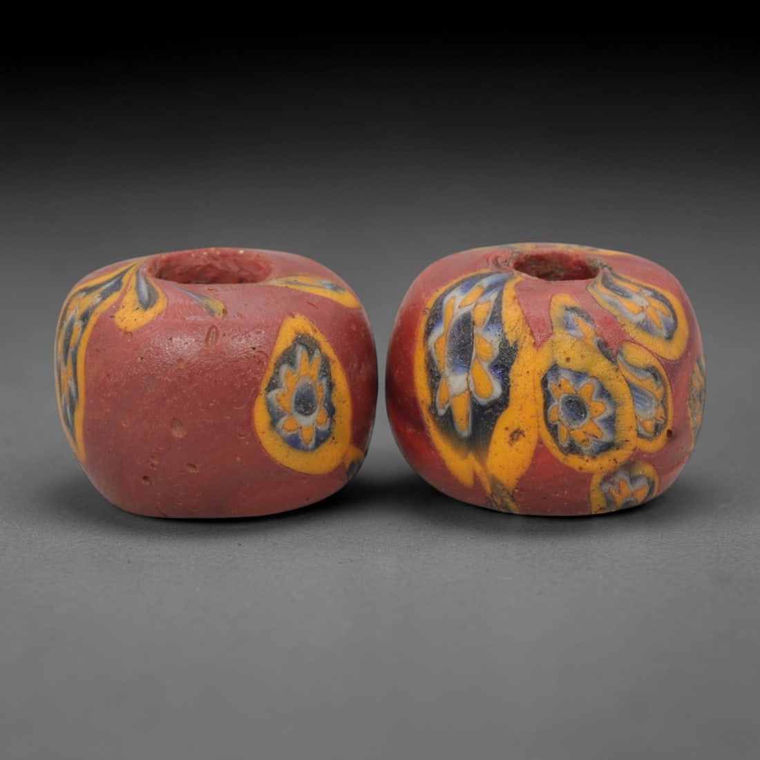 A Pair of Venetian Millefiori Glass Beads, 19th-20th Century, 50AD: Crafted in Venice during the 19th-20th century, this pair of beads is formed from opaque red glass, shaped into short cylinders with central drilled perforations. The surface is adorned with character