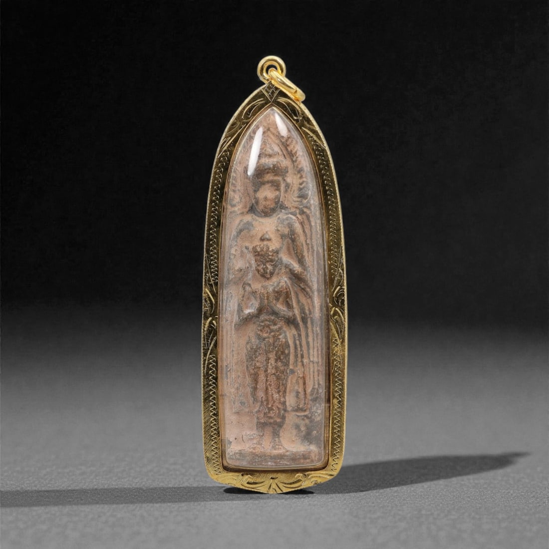 A Thai Buddhist Composite Amulet in Gilt Metal Frame, 20th Century, 57FN: The amulet is encased in an elongated, tapering gilt metal frame with a suspension loop at the apex and fine incised geometric border detailing. The obverse features a composite plaque of compressed o