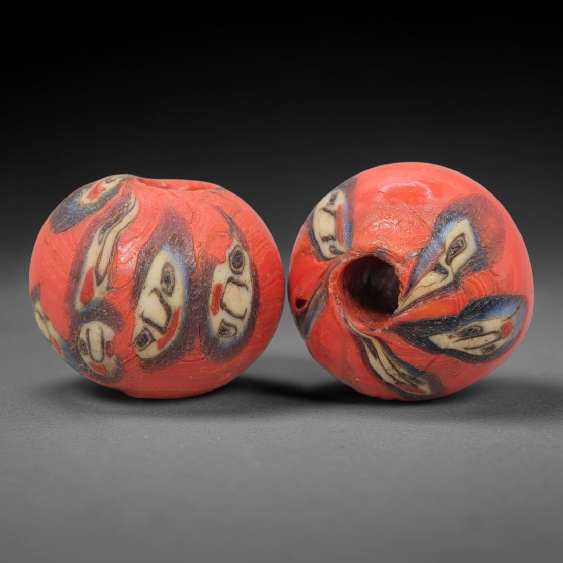 A Pair of Venetian Glass Trade Beads, 19th-20th Century, 24ZM: Formed as spherical beads, each is crafted from opaque red glass with a central perforation for suspension. The surface is decorated with multiple stylized human face motifs, featuring white, blue, an