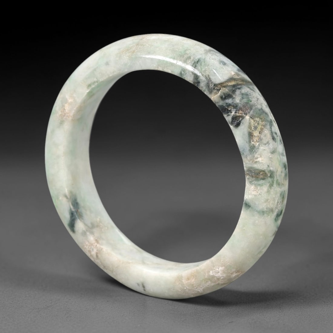 A Chinese Jadeite Jade Bangle Bracelet, 20th Century, 82ZK: Carved from jadeite jade with a pale greenish-white base and concentrated dark green, grey, and tan mineral inclusions on one section, the bangle is formed as a solid continuous circular band with a r