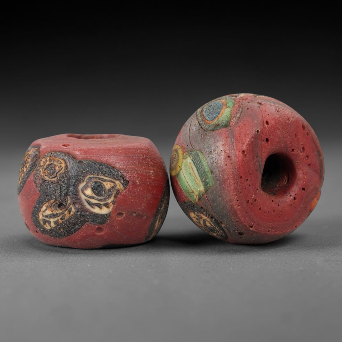 A Pair of Venetian Glass Trade Beads, 19th-20th Century, 88ZM: Formed as barrel-shaped beads, each is crafted from opaque red glass with a central perforation for suspension. The surface is decorated with multiple polychrome eye and abstract motifs, featuring dar