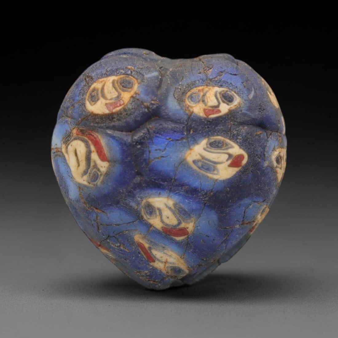A Venetian Mosaic Face Bead, 19th-20th Century, 59ZM: Fabricated from translucent blue glass, the bead is heart-shaped with a central perforation for suspension. The surface is densely decorated with multiple circular mosaic inlays depicting stylized hum
