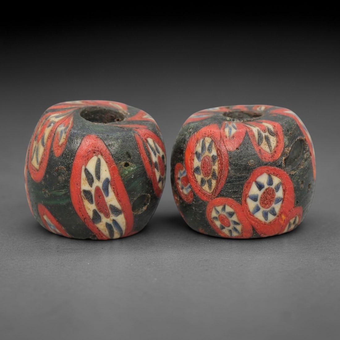 A Pair of Venetian Millefiori Glass Beads, 19th-20th Century, 62AD: Fabricated from opaque dark green glass, each bead is formed as a short cylinder with a central drilled perforation. The surface is decorated with characteristic millefiori canes, featuring circular e