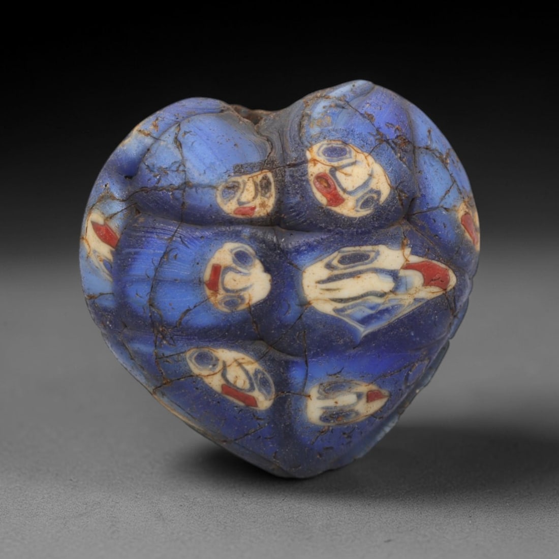A Venetian Mosaic Face Bead, 19th-20th Century, 60ZM: Fabricated from translucent blue glass, the bead is heart-shaped with a central perforation for suspension. The surface is densely decorated with multiple circular mosaic inlays depicting stylized hum