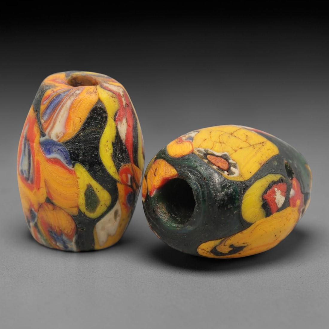A Pair of Venetian "End of Day" Glass Trade Beads, 19th-20th Century, 22ZM: Formed as elongated barrel-shaped beads, each is crafted from opaque glass with a central perforation for suspension, exemplifying the characteristic "End of Day" millefiori technique. The surface fea