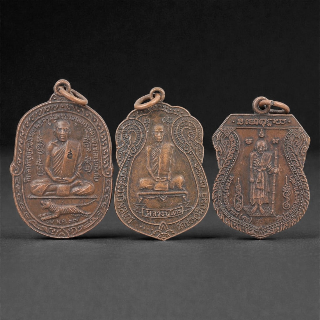 Three Thai Copper Alloy Devotional Amulets, 20th Century, 84FN: Cast from copper alloy with a rich brown patina, the group comprises three distinct devotional amulets, each with a suspension loop at the apex for wear. The first amulet is oval-shaped, depicting a s