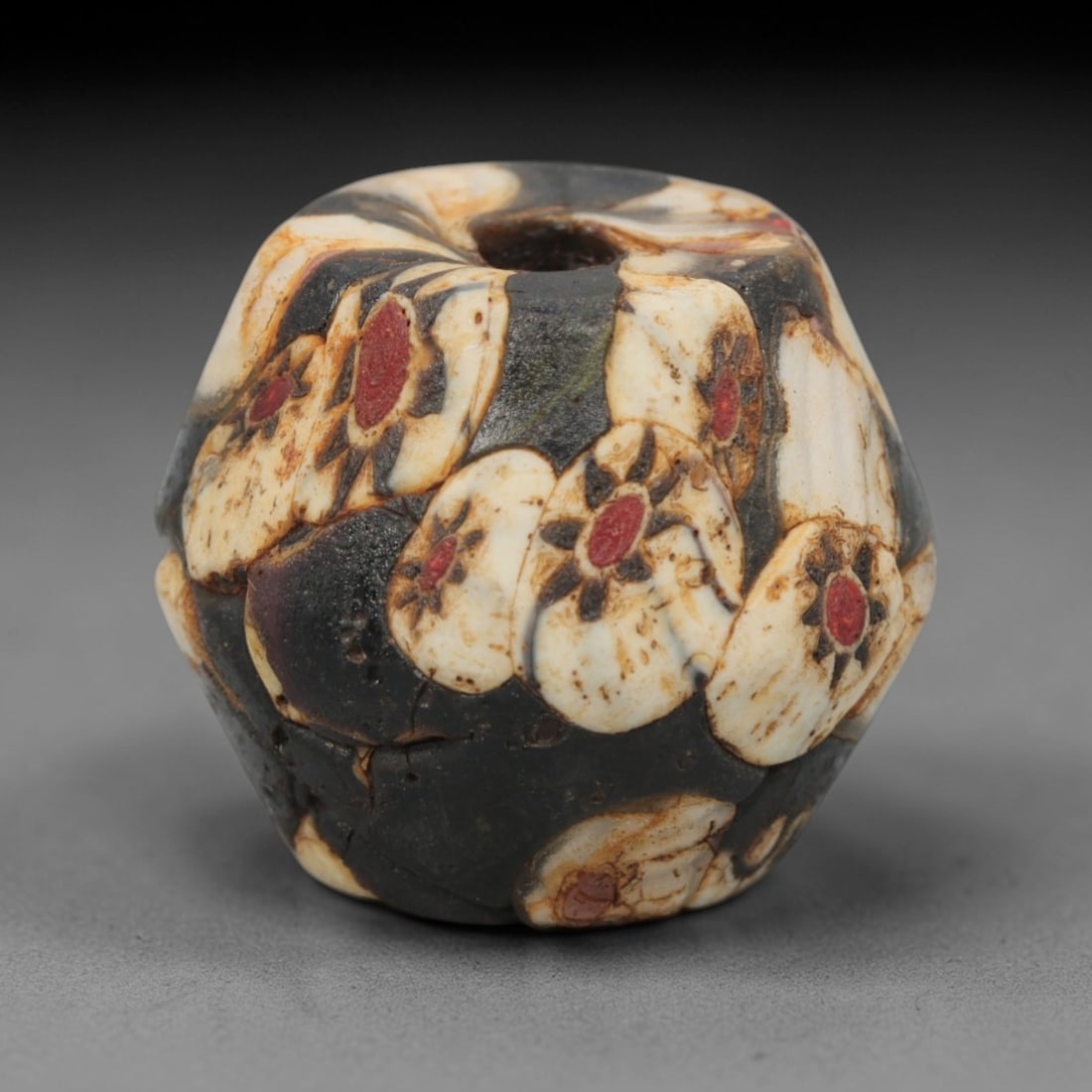 A Venetian Millefiori Glass Bead, 19th-20th Century, 75AD: Crafted in Venice during the 19th-20th century, this bead is formed from opaque dark glass, shaped into a faceted polyhedron with a central drilled perforation. The surface is adorned with characteris