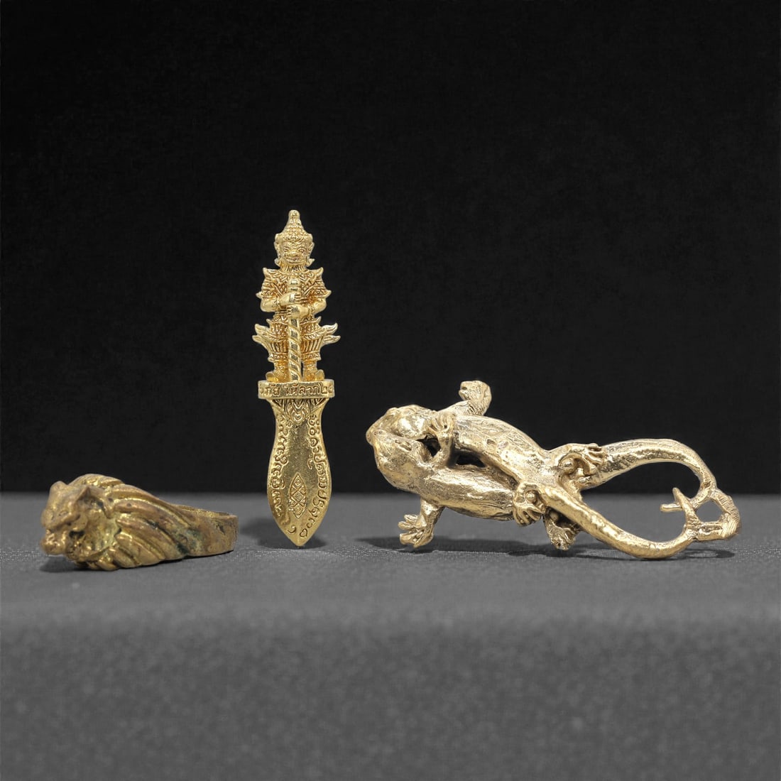 A Set of Three Thai Brass Amulet and Ornament Figures, 20th Century, 78ZG: Cast from brass with a warm golden patina, the set comprises three distinct objects: a ring, a dagger-shaped amulet, and a lizard-form ornament. The ring is shaped as a stylized beast head, with detai