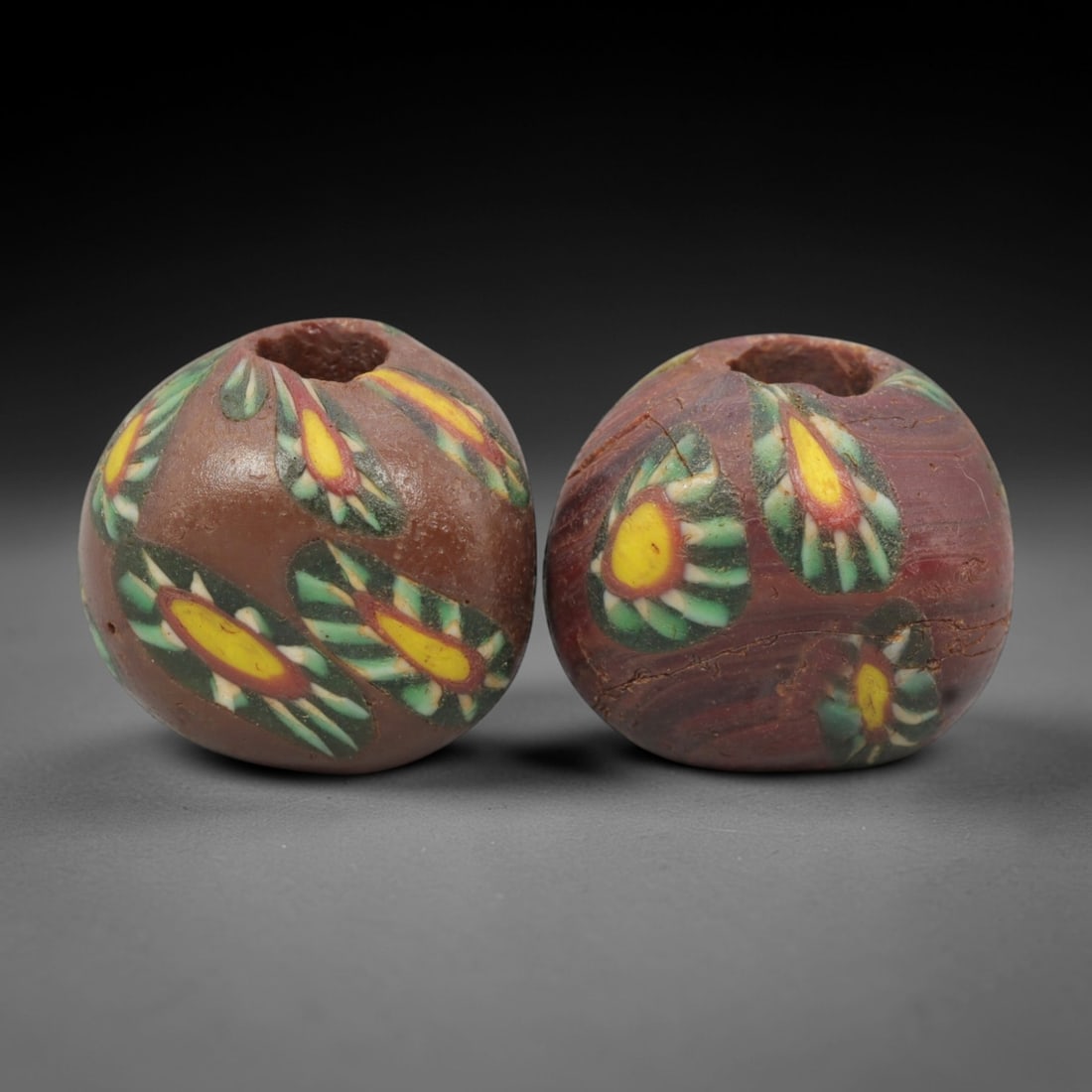 A Pair of Venetian Millefiori Glass Beads, 19th-20th Century, 43AD: Crafted from opaque brown glass, each bead is formed as a sphere with a central drilled perforation. The surface is decorated with characteristic millefiori canes, featuring eye-like motifs with yello