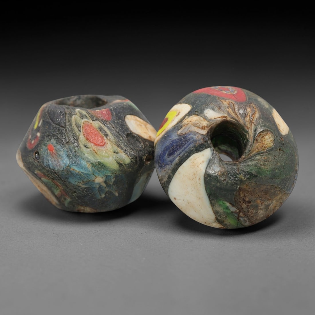 A Pair of Venetian Glass Trade Beads, 19th-20th Century, 16ZM: Formed as faceted and spherical beads, each is crafted from dark translucent glass with a central perforation for suspension. The surface displays embedded, fragmented inclusions of opaque colored gla